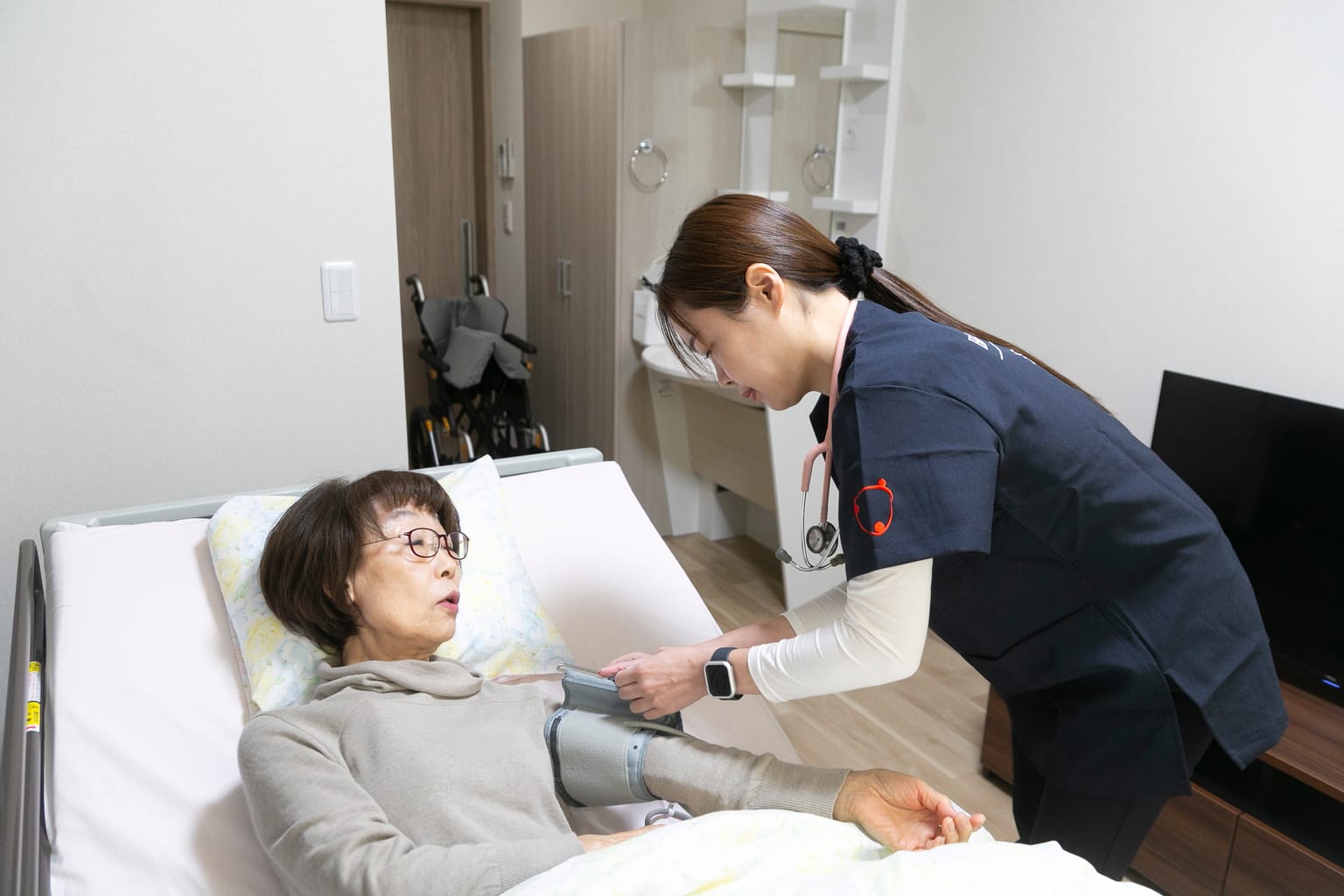 ベッドに横になった高齢の女性に血圧計を巻く訪問看護師(関連会社3SMILEの訪問看護の撮影より)
