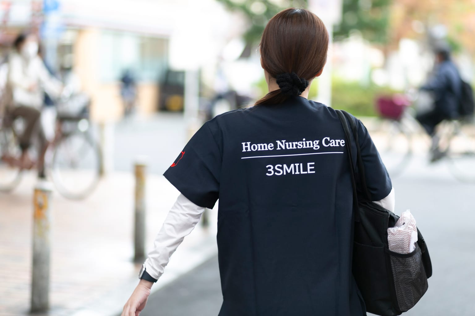 訪問看護の文字が入った制服の背中を見せて街を歩く訪問看護師(関連会社3SMILEの訪問看護の撮影より)
