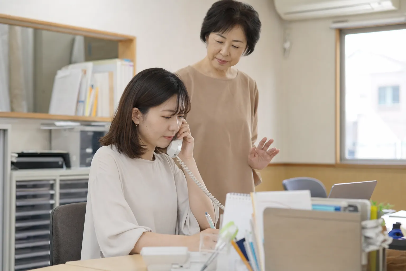 電話中の事務員の横で、先輩がそばに立って支えている(AIで生成したイメージ)