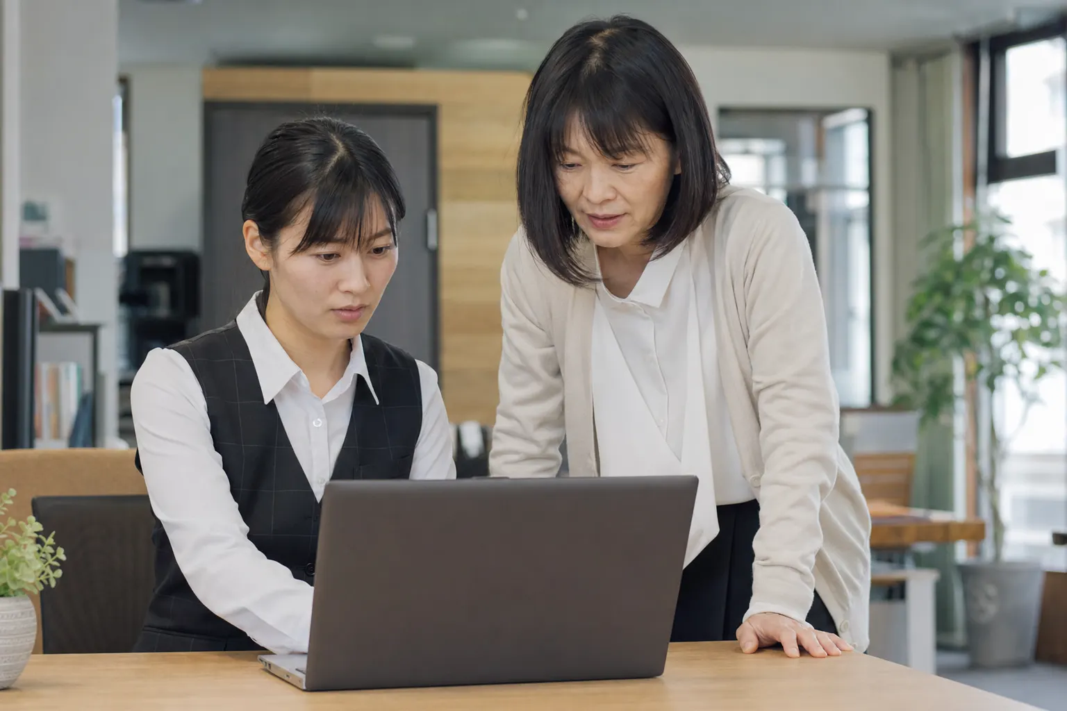 事務員がノートパソコンで作業し、横で同僚が確かめている(AIで生成したイメージ)